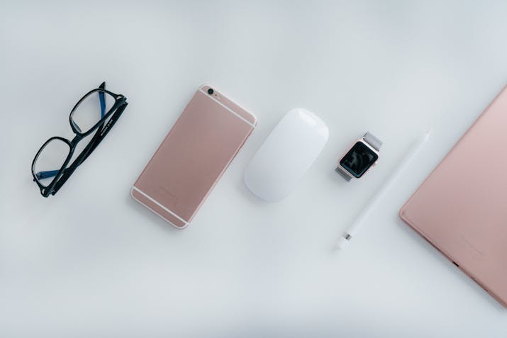Apple Watch Unpairing Explained 3 Flat lay of rose gold iPhone, Magic Mouse, Apple Watch, Apple Pencil, iPad, and glasses on white background.