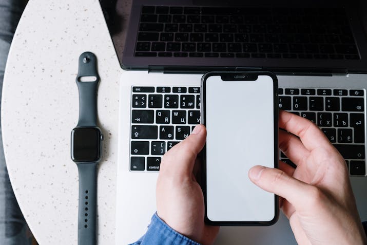 Apple Watch Unpairing Explained 1 Hands holding smartphone over laptop keyboard with Apple Watch on table.