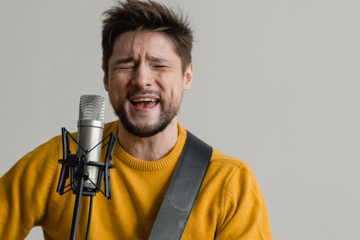 How to Avoid Choppy in GarageBand Playback 1 Bearded man in a mustard sweater singing into a studio microphone while playing guitar, eyes closed with expressive face against a plain background.