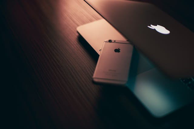 Apple’s iOS 26: A Major Leap in Design and Intelligence 1 A slightly open mac laptop with a iphone on its touch mouse pad in a dimly lit room on a dark colored table.